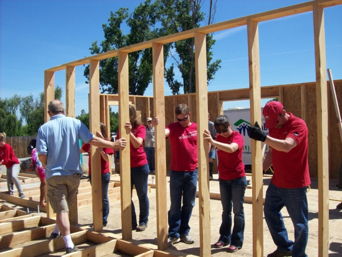 Volunteers constructing a wooden frame for a house under a clear blue sky