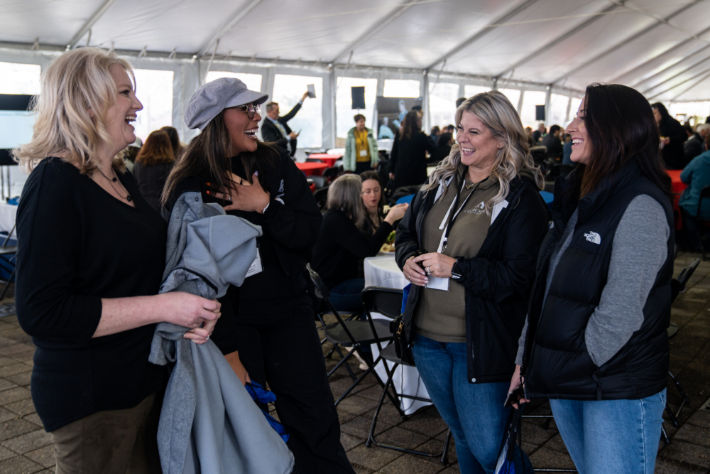 Four women laughing and chatting at an outdoor event under a large tent