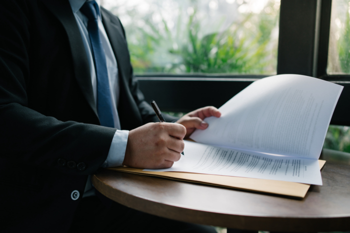Businessperson signing important documents at a round wooden table with natural light from a window.